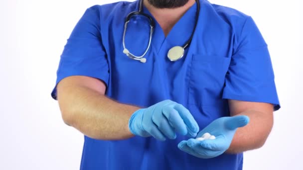 szakállas orvos férfi kék ruhában és kék gumikesztyű tartja fehér tablettát a kezében, és kínál egy fehér háttér, közelkép - Felvétel, videó