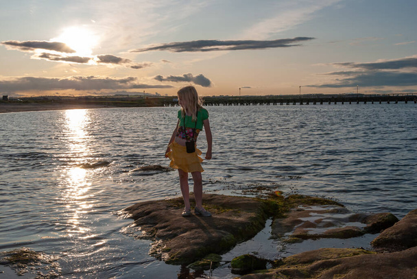 Menina ficou na rocha na borda da água com o pôr do sol atrás dela
 - Foto, Imagem
