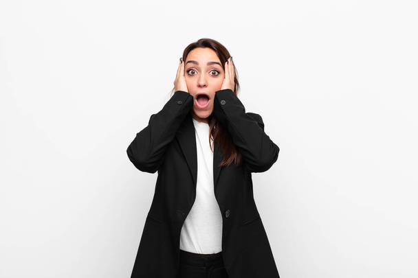 young pretty businesswoman looking unpleasantly shocked, scared or worried, mouth wide open and covering both ears with hands against white wall - Photo, Image