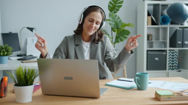 Happy businesswoman typing in office using laptop then dancing wearing headphones - Footage, Video