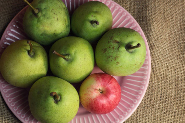 Zöld sárga organikus körte és egy kis vörös alma, más, mint a többi, finom rózsaszín tányéron, durva retro vásznon. Sötét hangulatú fotó gyümölcsökről, őszi októberi betakarítás felülnézet - Fotó, kép