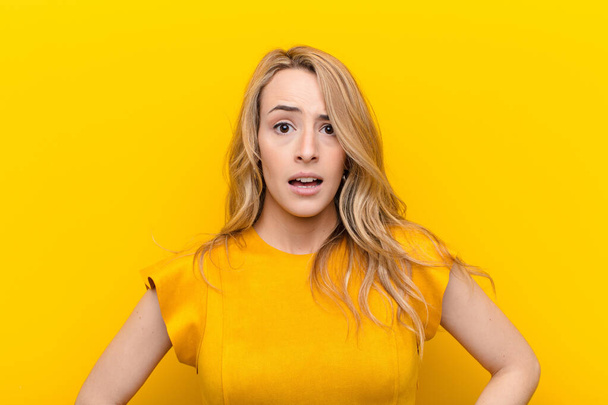 young pretty blonde woman looking very shocked or surprised, staring with open mouth saying wow against flat color wall - 写真・画像