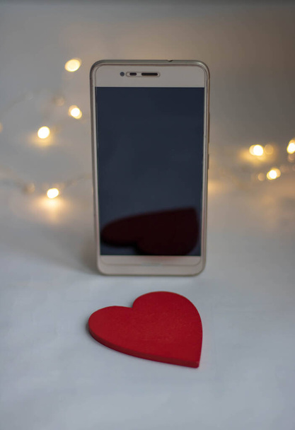 Телефон в подарок любимому человеку. Смартфон с красным бантом на светлом деревянном фоне с сердцем. Концепция дня Святого Валентина, любовь, сюрпризы. Плоская муха
 - Фото, изображение