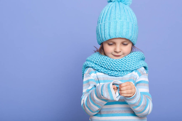 Picture of thoughtful cute little child, looking at her sleeve, wearing striped sweatshirt, blue scarf and hat, standing isolated over blue background in studio. Copyspace for advertisement. - Photo, image