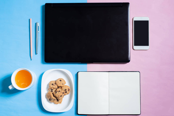 Ansicht von Laptop, Smartphone, Keks, Notizbuch, Stift, Bleistift, Orangensaft auf blauem und rosa Pastell - Foto, Bild