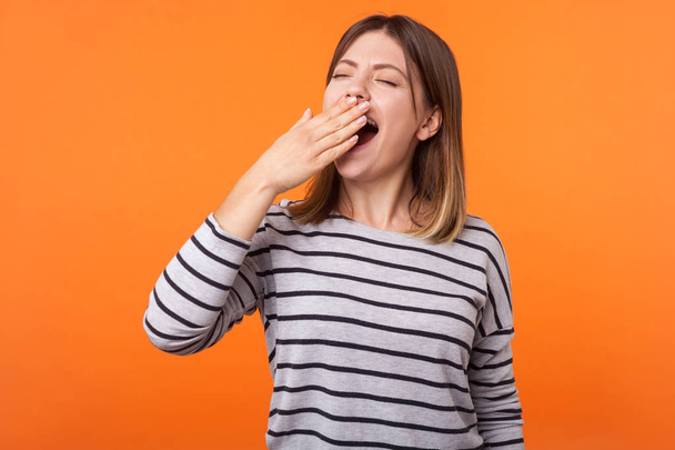 Portrait of exhausted or bored young woman with brown hair in long sleeve striped shirt standing, yawning and covering mouth with hand, disinterest. indoor studio shot isolated on orange background - Foto, immagini