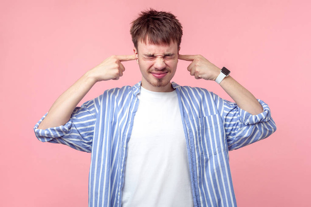 Don't want to hear! Portrait of annoyed brunette man with small beard and mustache in casual shirt standing with closed eyes, covering ears with fingers. indoor studio shot isolated on pink background - Foto, Bild