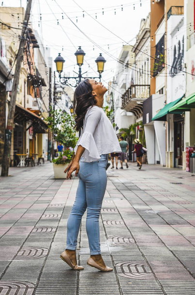 Outdoor portrait of young beautiful girl 9 to 25 years old posing in street. wearing white blouse and tight jeans and sapatillas. City lifestyle. Female fashion concept. - 写真・画像