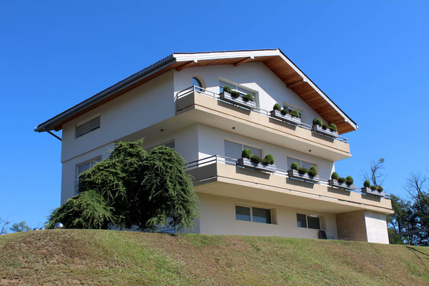 Grande casa di famiglia suburbana di nuova costruzione con lunghi balconi frontali sulla cima di una piccola collina circondata da erba appena tagliata e cielo blu chiaro sullo sfondo nella calda giornata estiva soleggiata
 - Foto, immagini