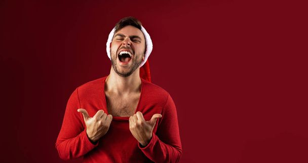 Young handsome caucasian guy in red sweater and Santa hats stands on red background in studio  Shows how hot he is - Zdjęcie, obraz