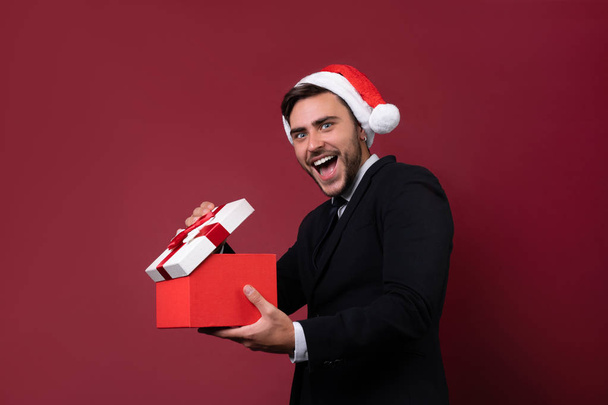 Young handsome caucasian guy in business suit and Santa hats stands on red background in studio and teeth smilie Holding red gift box in hand. - Фото, изображение