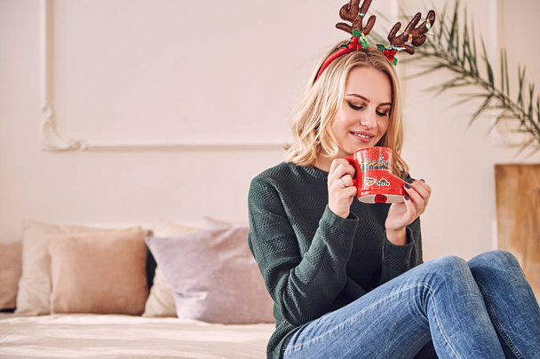 Menina bonita em orelhas de veado engraçado senta-se em um quarto acolhedor e segura uma xícara vermelha em suas mãos. Ambiente de Natal
. - Foto, Imagem