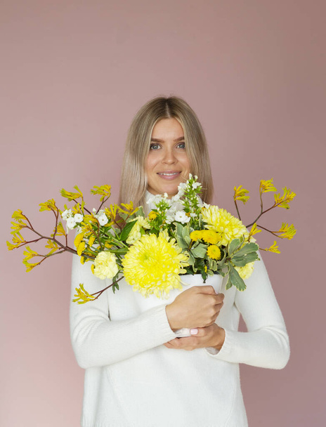 Een blonde mooie vrouw houdt in handen boeket van gele bloemen in een pot op een pastel roze studio achtergrond. Het concept van decoratie met bloemen, ikebana handgemaakte advertentie - Foto, afbeelding
