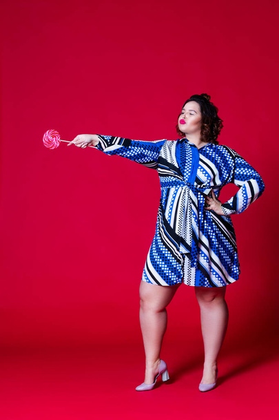 Plus size model in blue dress with big lollipop, fat woman on red background - Fotoğraf, Görsel