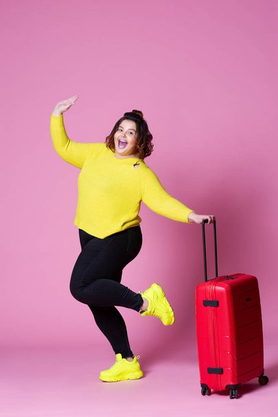 Cheerful plus size model with red suitcase, fat woman in black jeans and yellow jumper on pink background - 写真・画像