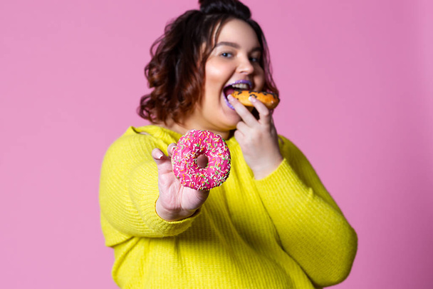 Χαρούμενη συν μέγεθος μοντέλο τρώει ντόνατς, η χοντρή γυναίκα overeats σε ροζ φόντο - Φωτογραφία, εικόνα