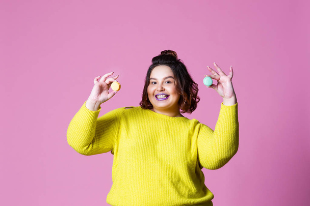 Cheerful plus size model posing with macaroon, fat girl loves sweets, pink studio background - Foto, Bild