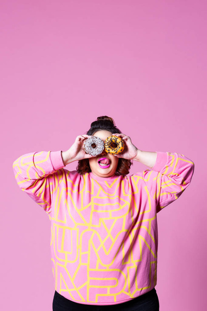 Cheerful plus size model holding donuts against her eyes and licks lips on pink background, body positive concept with copy space - Valokuva, kuva