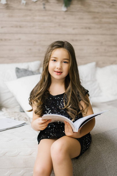 Dívka šest let bez dvou horní mléčné zuby v černé ženy drží v rukou knihy nebo časopisu, čte jeho, sedí na útulné posteli a usmívá se - Fotografie, Obrázek