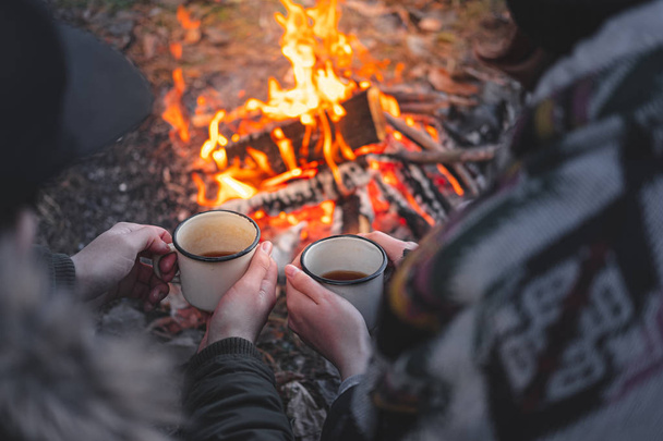 Two people warming hands with hot drinks by the bonfire. - Фото, изображение