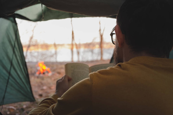L'uomo siede in tenda vicino al falò e legge un libro
.  - Foto, immagini