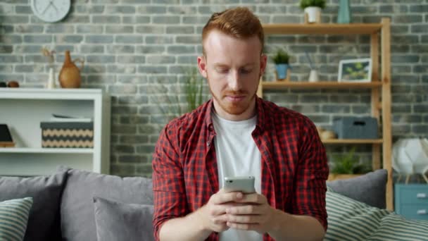 Red-haired bearded guy texting with smartphone smiling in cozy apartment - Záběry, video