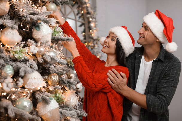 Happy young couple in Santa hats decorating Christmas tree toget - Foto, imagen