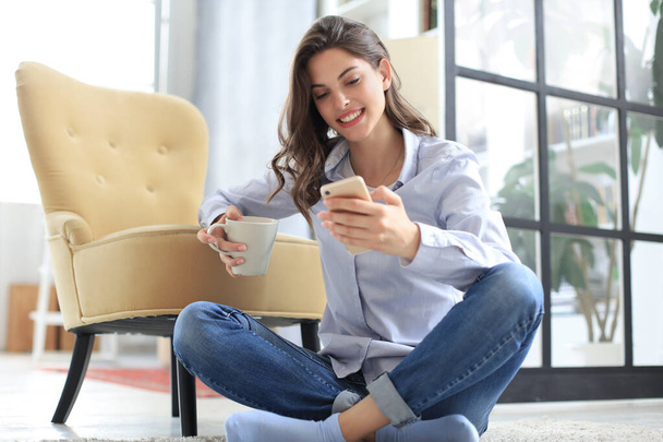 Smiling young female sitting on the floor in the living room and using with her mobile phone. - Foto, Bild