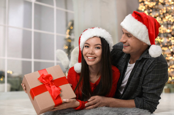 Happy young couple in Santa hats with Christmas gift at home - Фото, зображення