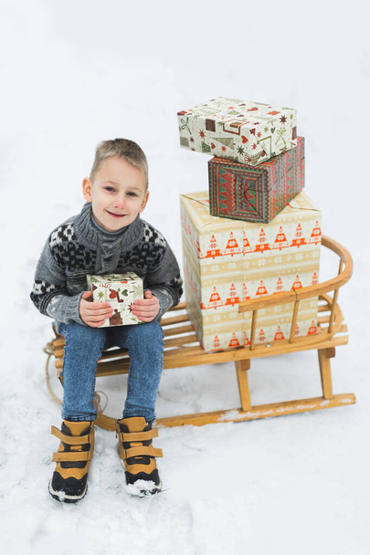 Little boy in gray knitted sweater, sitting on the wooden sledge, decorated with beautiful boxes with Chrristmas presents. Winter time, outdoors in winter forest or park. Child play in snowy park - Zdjęcie, obraz