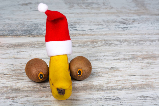 Banana y kiwi para el nuevo año. Plátano en un sombrero de Navidad y dos kiwi con ojos sobre un fondo de madera blanca. Vista frontal, lugar para texto, espacio para copiar
. - Foto, Imagen