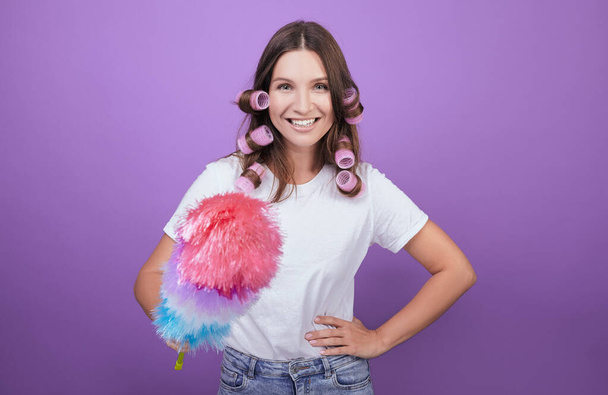 Cheerful girl in curlers removes dust with a brush. - Фото, изображение