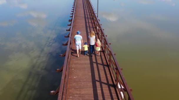 ευτυχισμένη οικογένεια Καυκάσιος κρατώντας τα χέρια περπατώντας ξύλινη γέφυρα πάνω από το νερό της λίμνης - Πλάνα, βίντεο