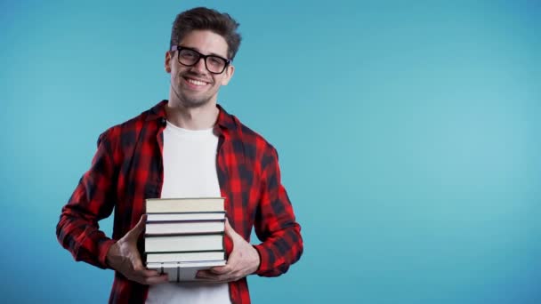 European student in red plaid shirt on blue background in studio holds stack of university books from library. Copy space. Guy smiles, he is happy to graduate. - Video