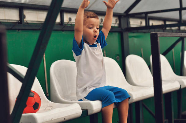 Pieni kolmevuotias poika sinisessä jalkapallounivormussa ja punaisella jalkapallollaan.
 - Valokuva, kuva