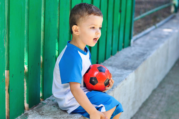 Petit garçon de trois ans en uniforme de football bleu et avec son ballon de football rouge
 - Photo, image
