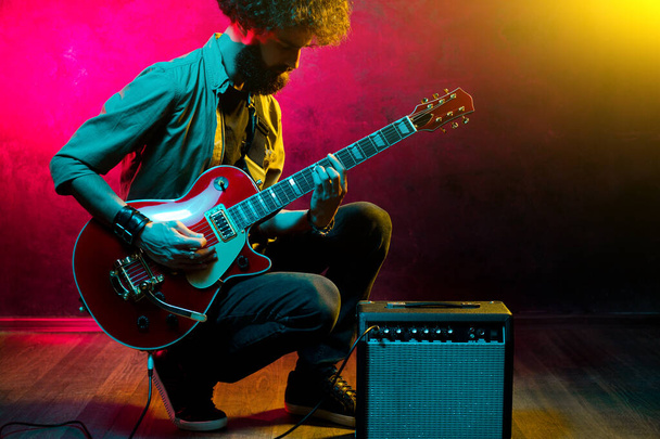 Portrait of hipster man with curly hair with red guitar in neon lights. Rock musician is playing electrical guitar. - Foto, Imagem