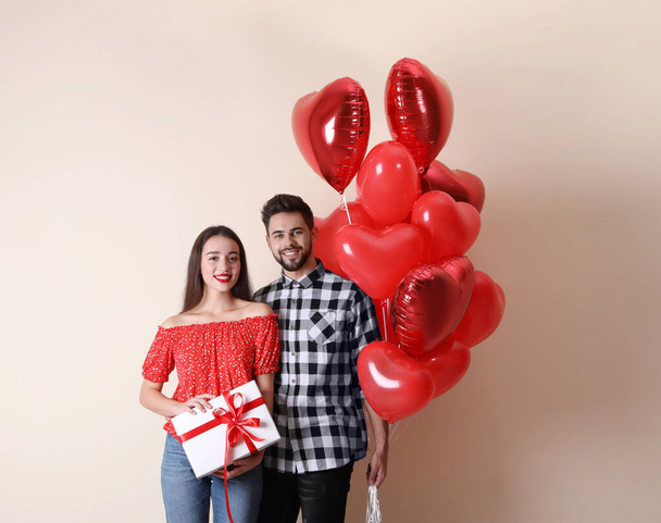 Happy young couple with gift box and heart shaped balloons on beige background. Valentine's day celebration - Foto, Imagen