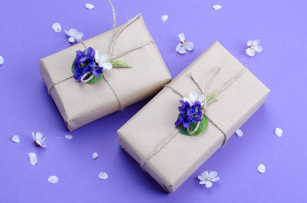 Красивые подарочные коробки, завернутые в простую коричневую бумагу, украшенную живыми цветами фиолетового цвета на фиолетовом фоне
. - Фото, изображение