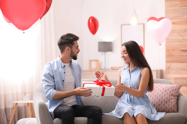 Giovane uomo che presenta regalo alla sua ragazza in soggiorno decorato con palloncini a forma di cuore. Festa di San Valentino
 - Foto, immagini