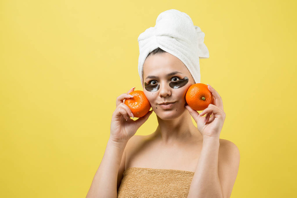 Beauty portrait of woman in white towel on head with gold nourishing mask on face. Skincare cleansing eco organic cosmetic spa relax concept. A girl stands with her back holding an orange mandarin. - Photo, Image