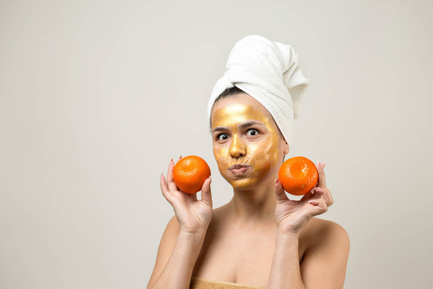 Beauty portrait of woman in white towel on head with gold nourishing mask on face. Skincare cleansing eco organic cosmetic spa relax concept. A girl stands with her back holding an orange mandarin. - Фото, изображение