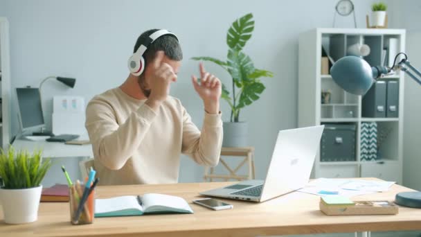 Cheerful office worker wearing headphones moving arms dancing and using laptop - Πλάνα, βίντεο