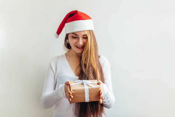 Mooi meisje met lang haar in rood Santa Claus hoed houden geschenkdoos geïsoleerd op witte achtergrond op zoek gelukkig en opgewonden. Jonge vrouw portretteert echte emoties. Gelukkig kerstfeest en nieuwjaarsvakantie - Foto, afbeelding