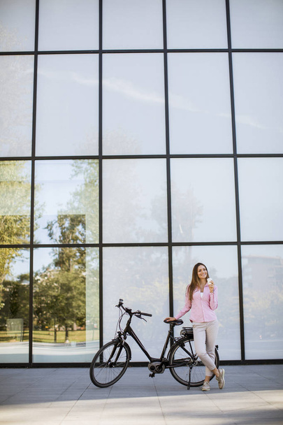 Красивая молодая велосипедистка пьет кофе из чашки на электрическом велосипеде в городской среде
 - Фото, изображение