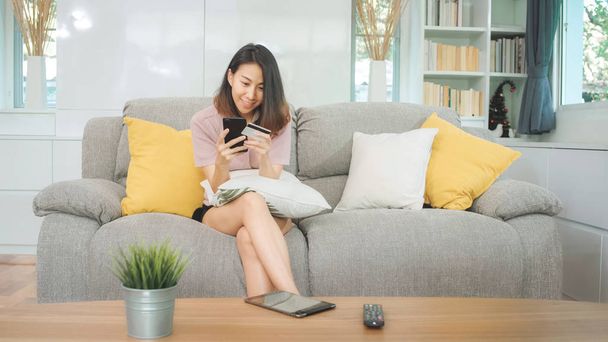 Young smiling Asian woman using smartphone buying online shopping by credit card while lying on sofa when relax in living room at home. Lifestyle latin and hispanic ethnicity women at house concept. - Foto, imagen