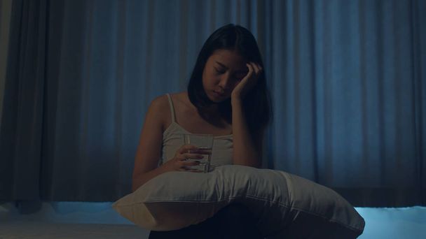 Kaunis aasialainen nuori nainen istuu sängyllä ottaa unilääkettä tai yölääkettä makuuhuoneessa. Epäterveellinen sairas intialainen nainen kärsii unettomuudesta tai päänsärystä, masentunut tyttö pitää masennuslääkkeitä. - Valokuva, kuva