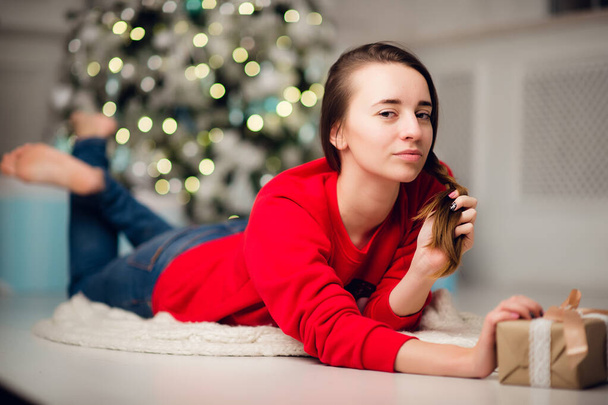 holidays, celebration and people concept - smiling woman wearing red sweather and jeans holding gold gift box over christmas tree lights background - Foto, imagen