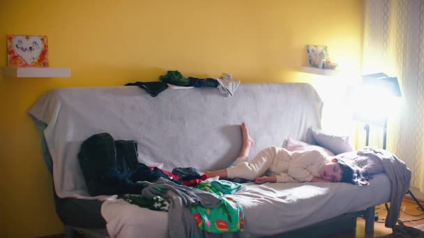 Το αγόρι κοιμάται στον καναπέ στο σπίτι. Γύρω από σκόρπια ρούχα. - Πλάνα, βίντεο