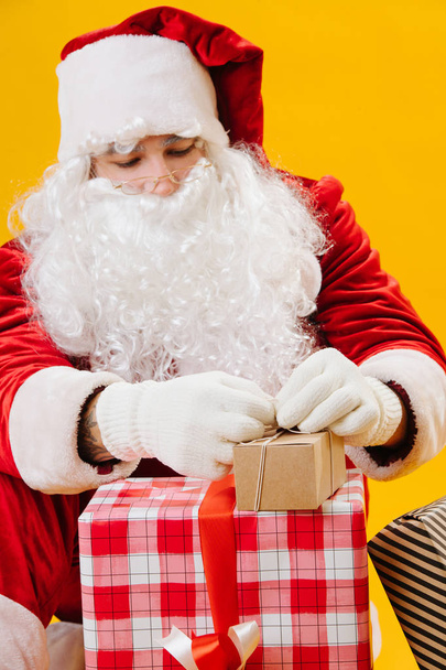 Weihnachtsmann sitzt auf dem Boden und bindet vorsichtig kleine Geschenke vor sich her. - Foto, Bild